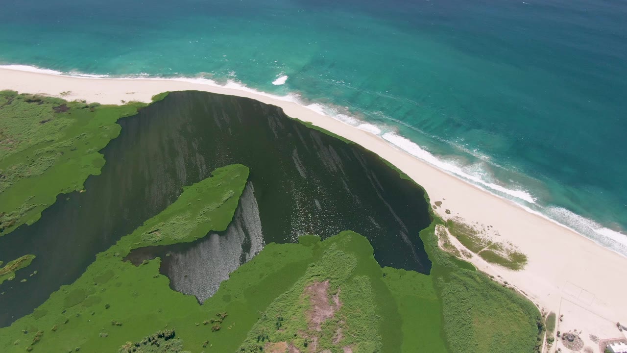 San Jose del Cabo aerial frame with the grassy bluff centered, a dark cliff wall below it, white beach at right, and no visible people.