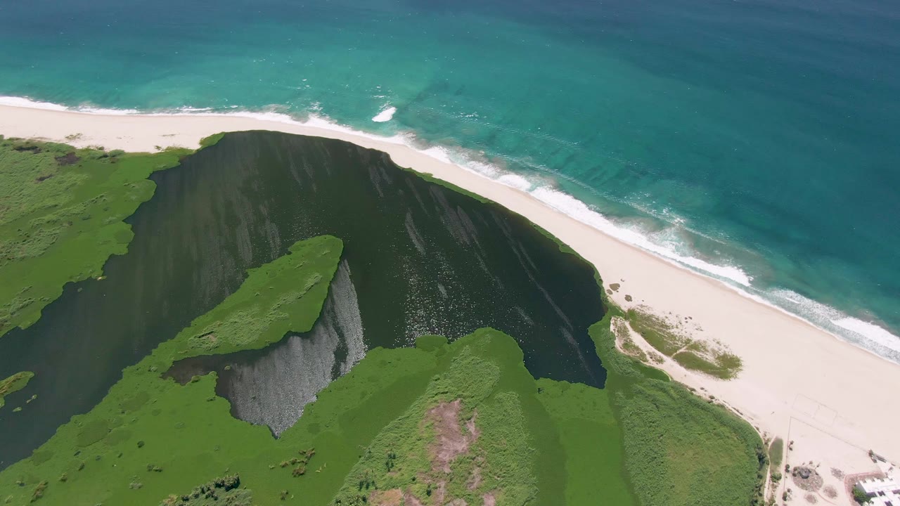 Aerial view over San Jose del Cabo coastline with steep green terrain, a pale beach curving along the water, and no visible people.