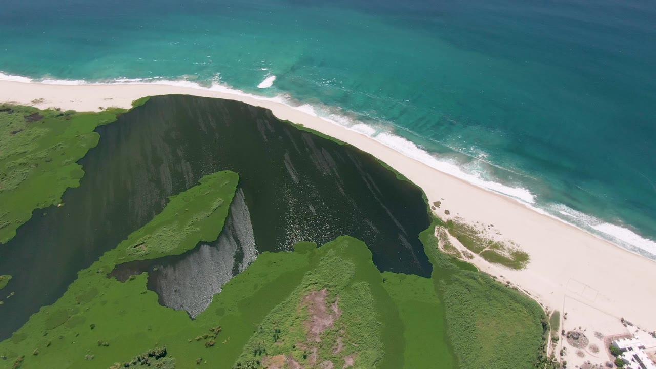 San Jose del Cabo coastline from above, with the dark cliff face shifted left, broad white sand at right, rolling surf, and no visible people.