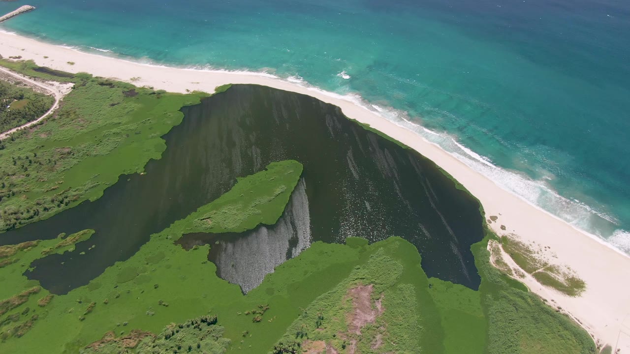 Overhead San Jose Del Cabo coastline with the green cliff centered, dark lagoon water around it, white sand across the upper frame, and no visible people.