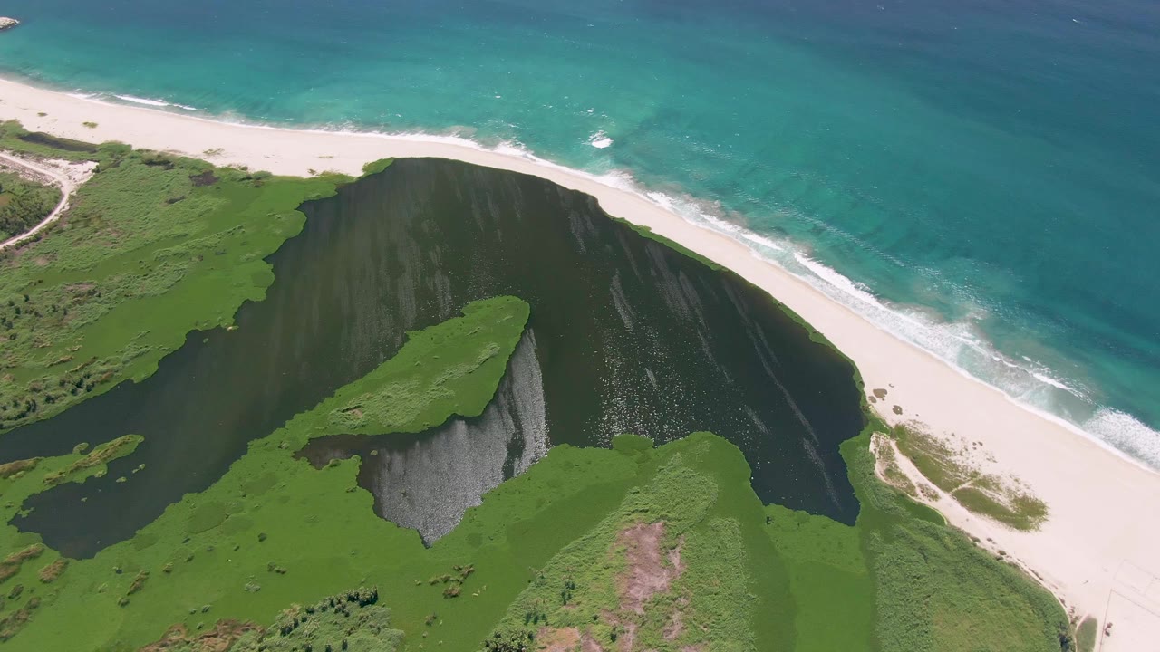 Aerial frame from San Jose Del Cabo showing the lagoon wrapping around the green cliff, the sandy shore above it, turquoise surf, and no visible people.