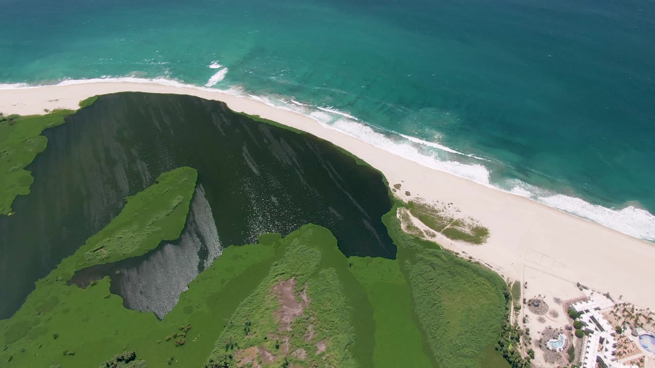 At 0:01, the San Jose Del Cabo aerial view holds on a dark green cliff face, pale beach, foamy shore break, turquoise water, and a small resort edge with no visible people.