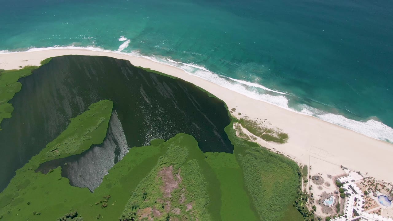 At 0:02, the aerial composition in San Jose Del Cabo includes green cliffs in the lower frame, a clean curve of beach, white surf lines, blue-green ocean, and no visible people.
