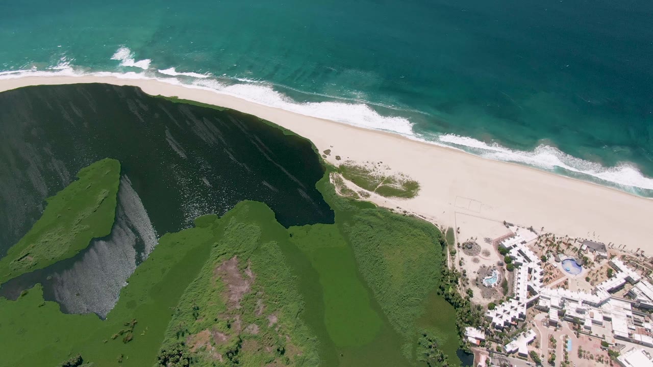 Aerial view in San Jose del Cabo showing turquoise ocean surf above a pale sandy beach, steep green hills in the foreground, white buildings at lower right, and no visible people.