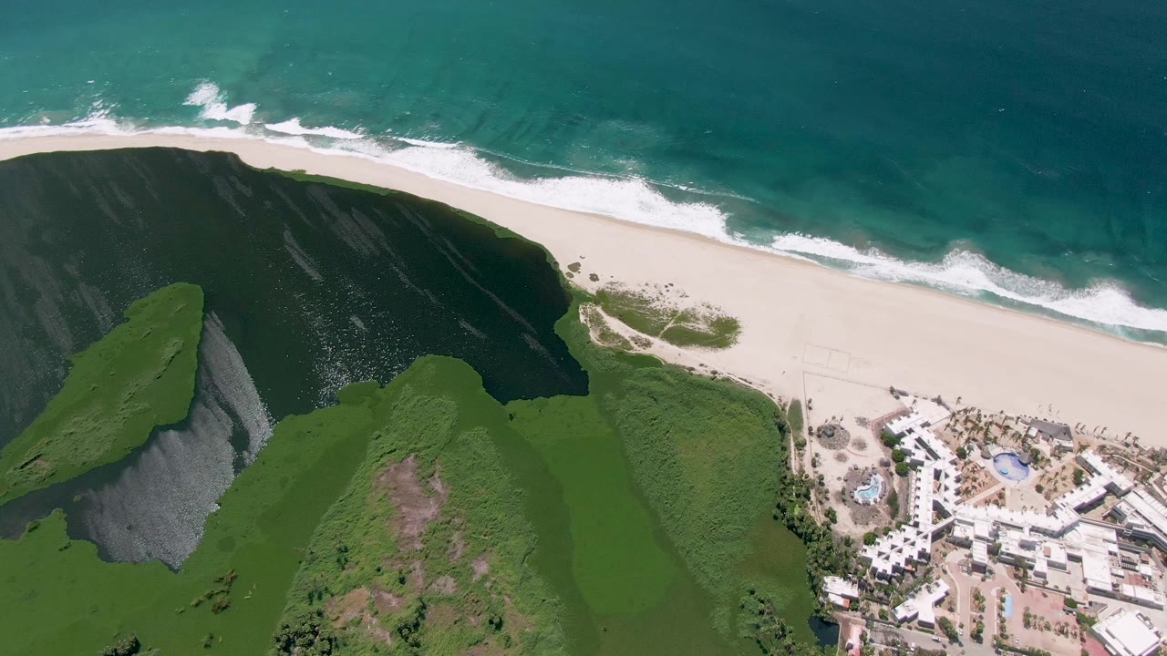 Overhead coastal frame with waves, sandy shoreline, green hillside, and resort buildings near the lower right.