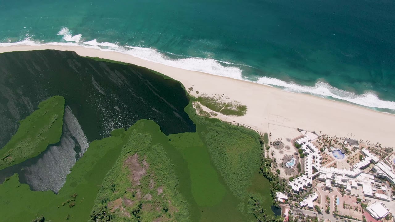 At 0:02, the San Jose del Cabo coastline remains in overhead view, with foamy surf rolling onto sand, green hillside filling the lower frame, buildings at the right edge, and no visible people.