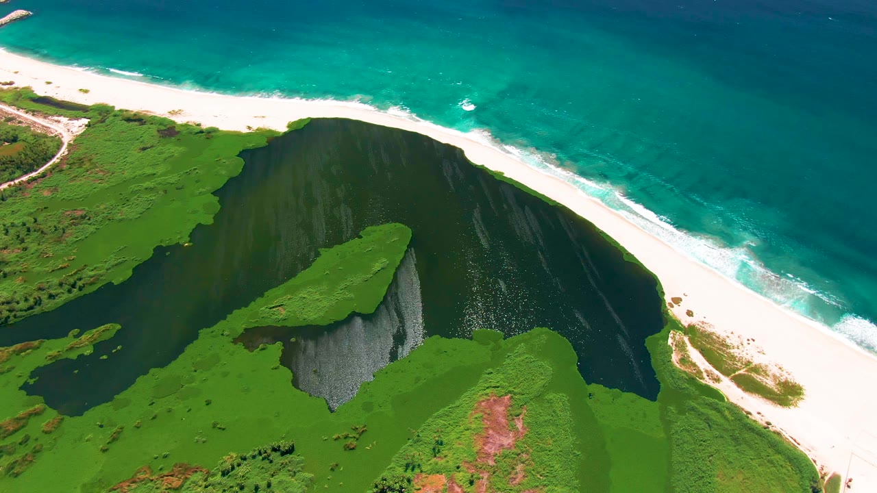 Aerial frame from San Jose Del Cabo showing green cliff tops around a dark inlet, a diagonal white sand beach, turquoise ocean water, and no visible people.