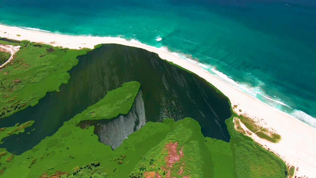 San Jose Del Cabo aerial frame with a green bluff projecting into dark coastal water, bright sand above it, turquoise sea beyond, and no visible people.