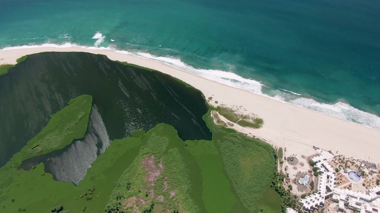 Aerial frame from San Jose Del Cabo showing turquoise water, a pale beach, green cliff tops, shadowed slopes, and no visible people at the start of the clip.