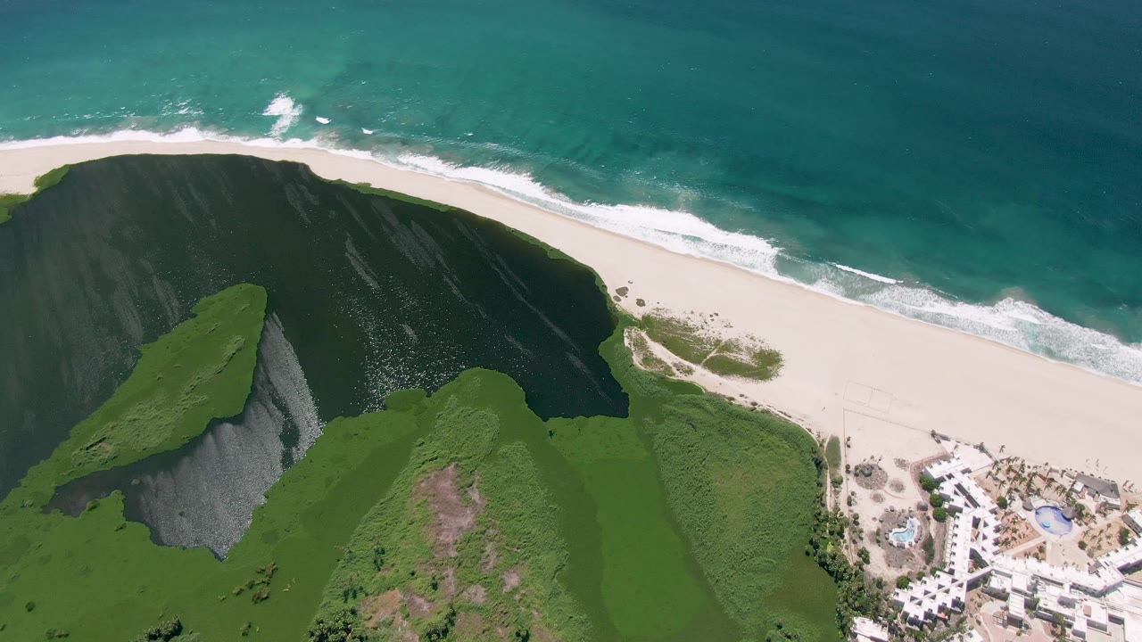 Aerial San Jose Del Cabo coastline view with white surf bands closer to the sand, green cliffs below, turquoise ocean above, and no visible people.