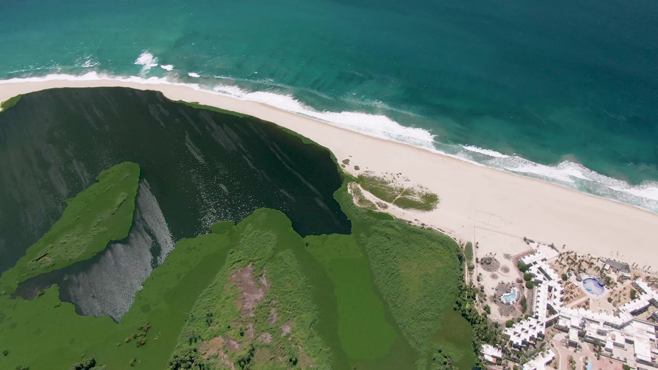 Aerial shoreline with broad cliff shadow, bright sand, and rolling surf.