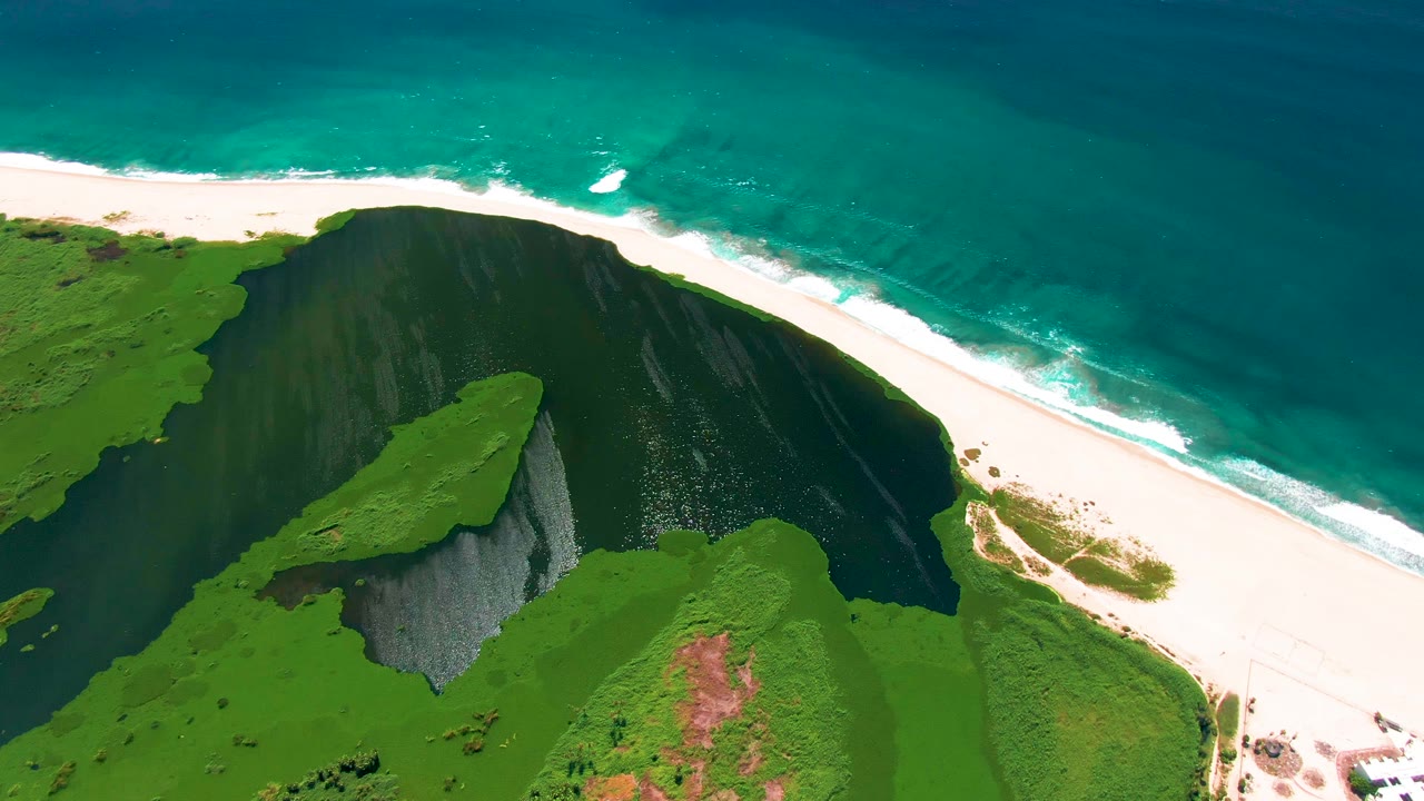 Aerial view in San Jose Del Cabo, Baja California Sur, showing green cliffs casting dark shadow toward a white sand beach and turquoise ocean, with no visible people.