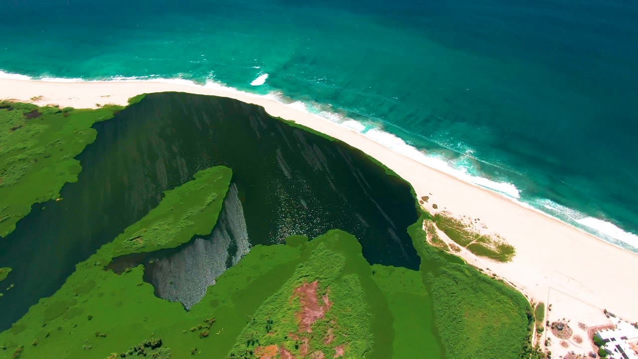 Green coastal bluff, pale beach, and blue-green water seen from above.