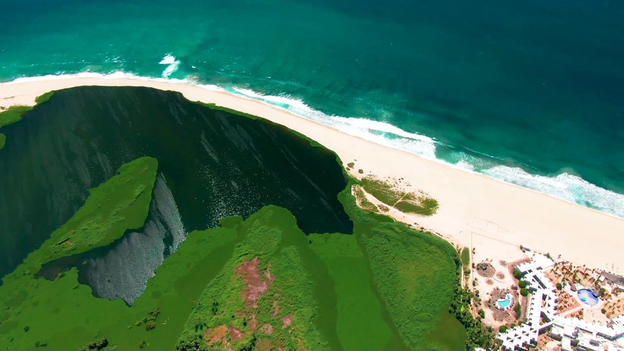 Aerial view in San Jose del Cabo with the green cliff and dark cliff face prominent, waves breaking along the white sand, resort structures at lower right, and no visible people.