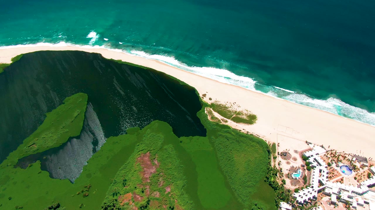 Aerial frame over San Jose del Cabo with the cliff shifted lower in the composition, surf tracing the beach, turquoise water above, resort buildings at lower right, and no visible people.