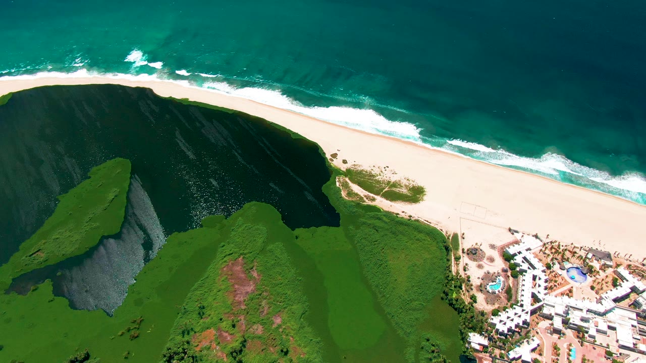 Aerial frame over San Jose del Cabo showing turquoise ocean at the top, white surf on a wide sandy beach, green cliff slopes below, and white resort buildings with pools at the lower right, with no visible people.