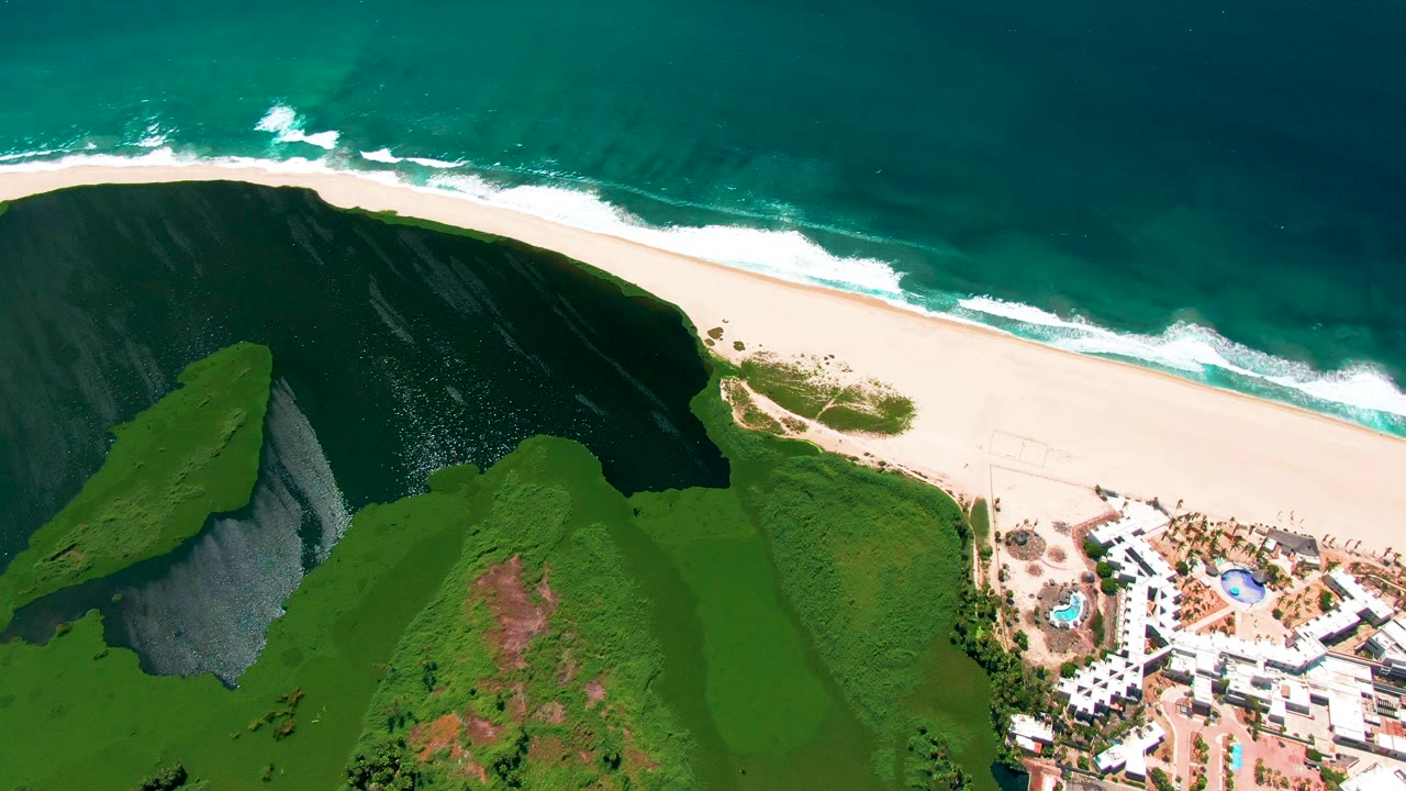 Aerial view in San Jose del Cabo with the surf line slightly farther along the beach, green cliff terrain filling the lower half, and the white resort complex and blue pools visible near the lower right, with no visible people.