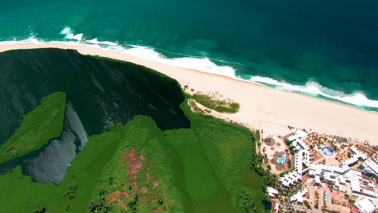 Aerial beach scene with ocean waves, steep green cliff, and resort buildings.