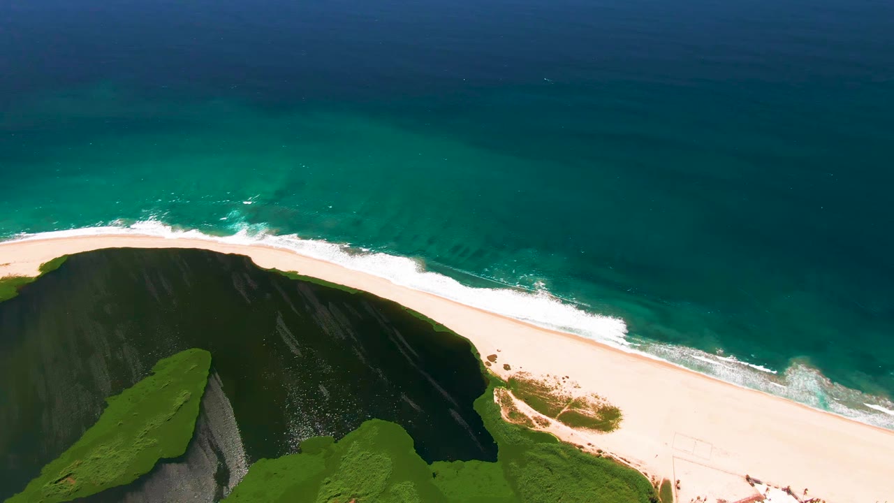 High aerial view over San Jose Del Cabo coastline with green cliff vegetation at the lower edge, a pale sandy beach, turquoise ocean, white surf, and no visible people.
