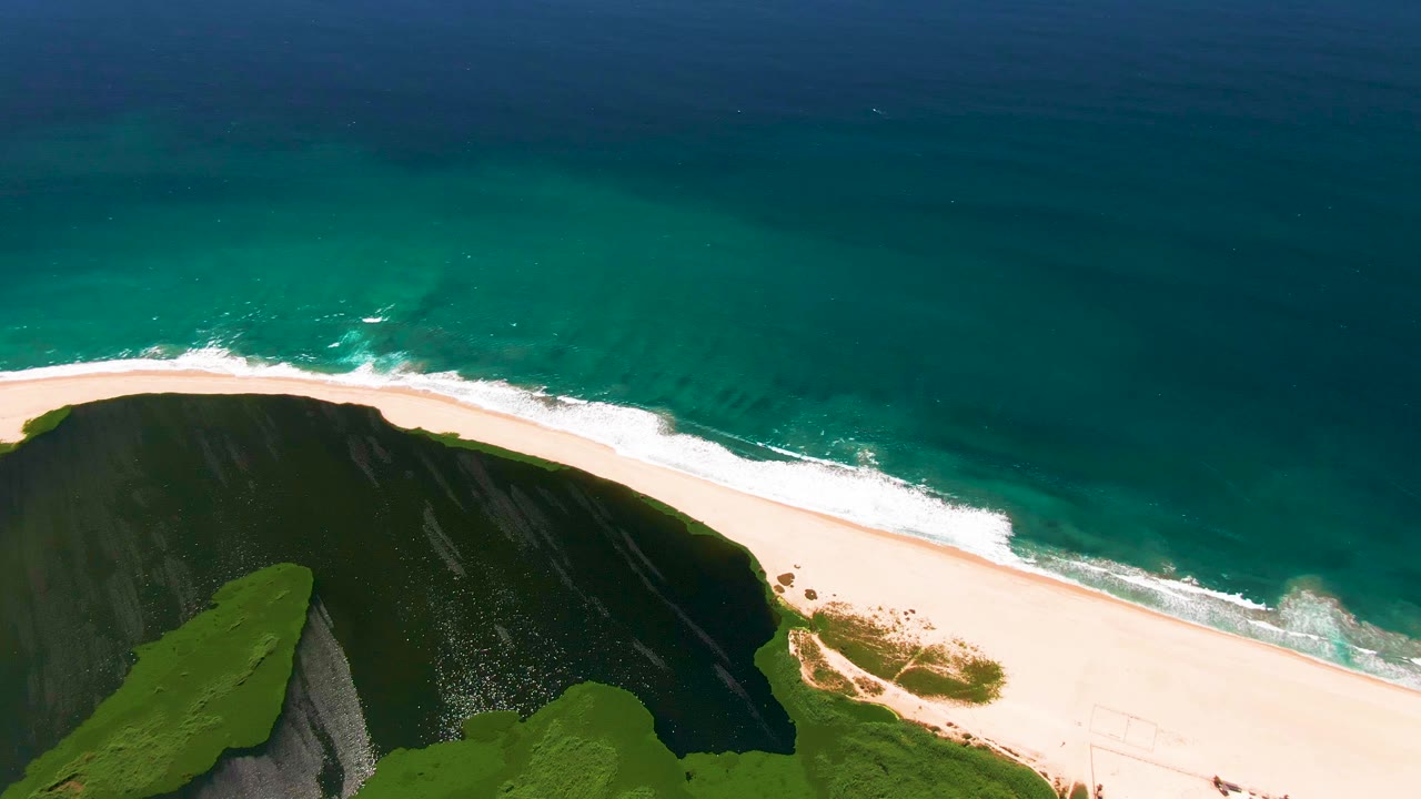 Aerial San Jose Del Cabo frame with the beach stretching diagonally across the image, dark cliff below, green vegetation in the corner, turquoise water above, and no visible people.