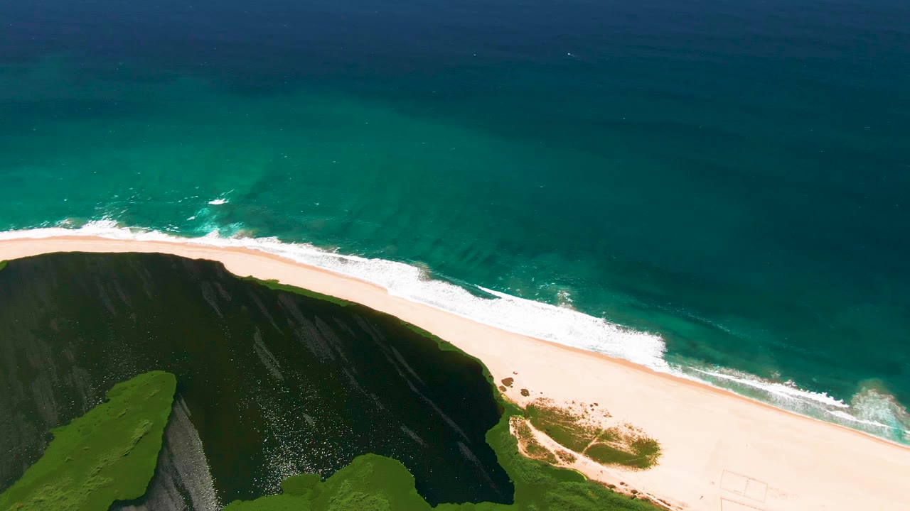 High aerial view in San Jose Del Cabo with a longer sweep of pale beach, small patches of dune vegetation near the sand, white surf along the edge, and no visible people.