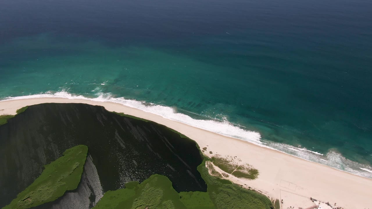 Aerial view of a green cliff above a sandy beach and turquoise ocean.