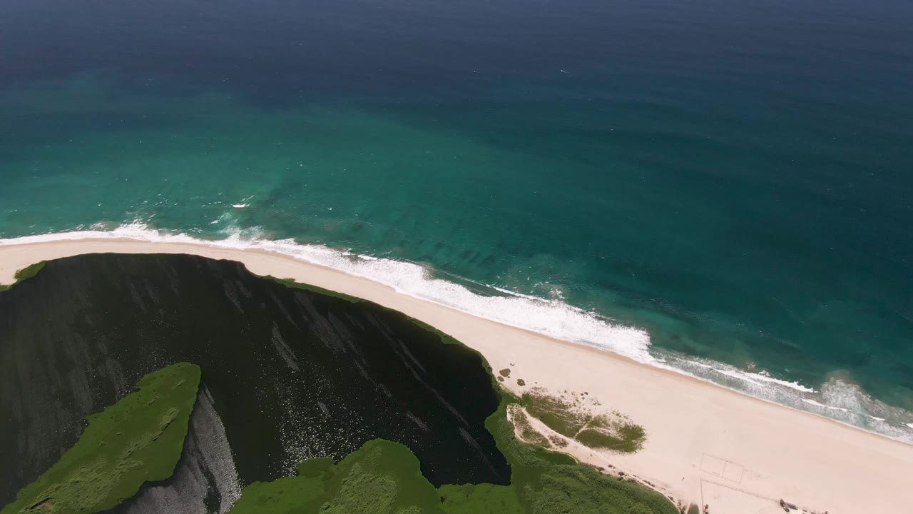 At 0:01, the San Jose del Cabo shoreline remains framed from above with the grassy cliff lower left, white surf spreading along the sand, and no visible people.