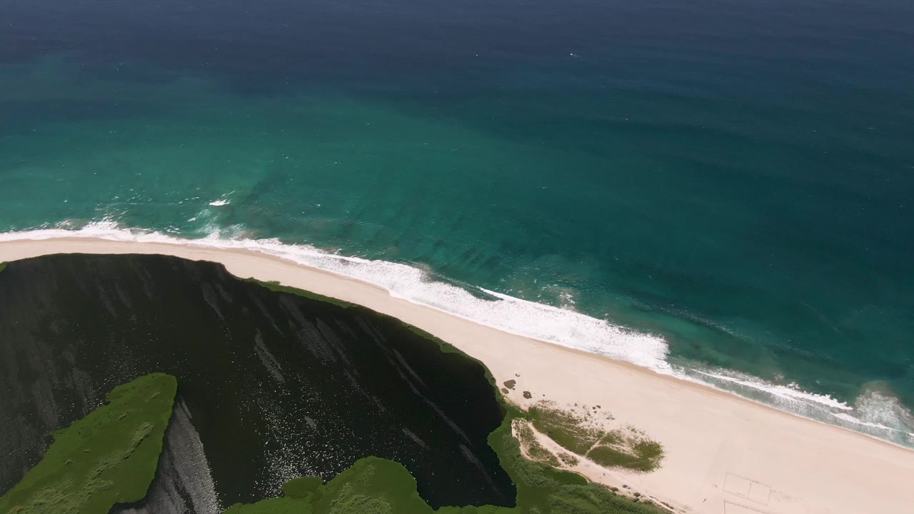 At 0:02, the aerial view over San Jose del Cabo shows the cliff point lower left, a pale beach curving across frame, foamy surf, and open blue water with no visible people.