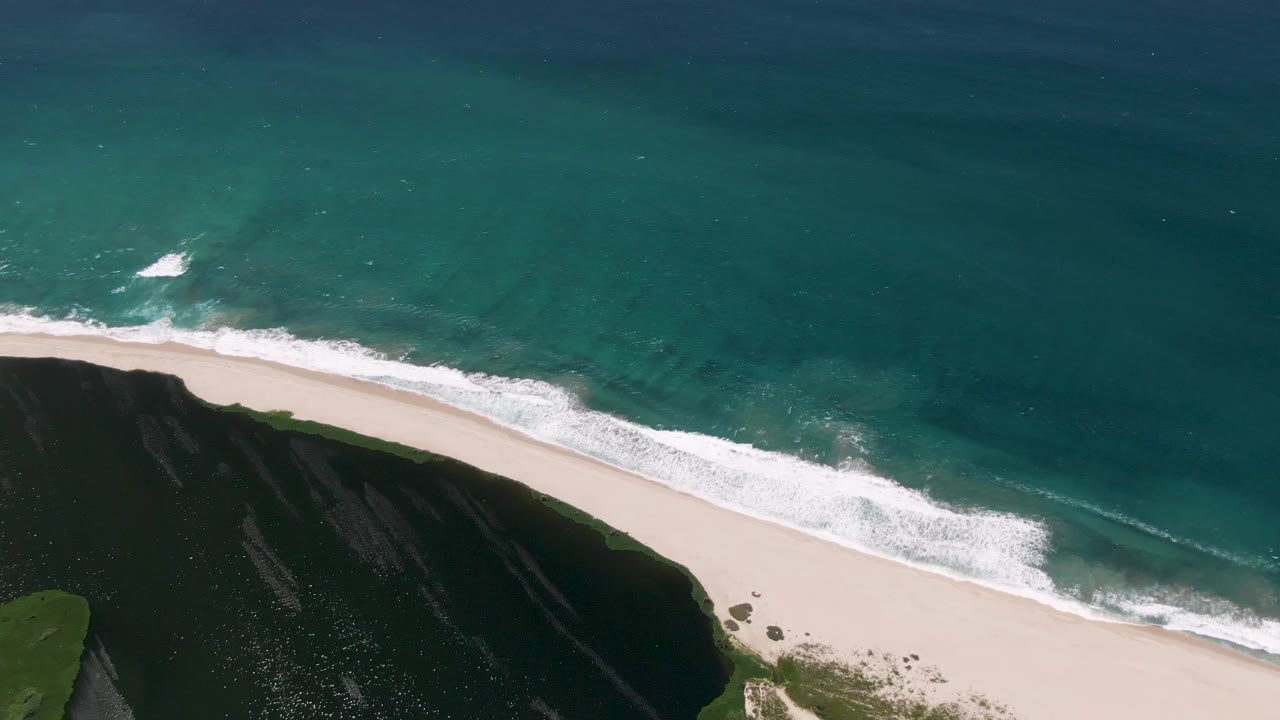 Aerial frame from San Jose del Cabo showing turquoise water above a pale sandy beach, white surf near shore, a dark cliff edge at lower left, and no visible people.