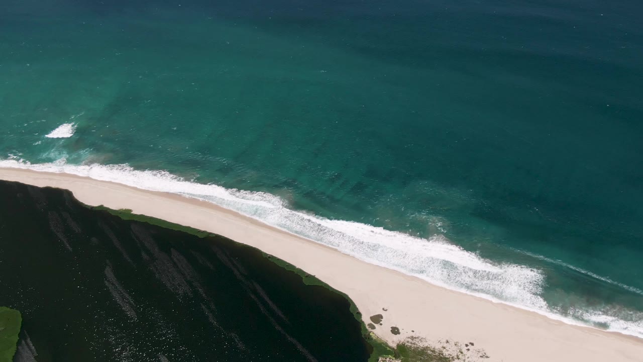 Aerial San Jose del Cabo beach view with a longer band of white wave foam crossing the center, turquoise ocean above, dark green cliff at lower left, and no visible people.