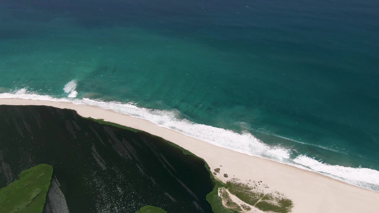 Aerial frame over San Jose del Cabo coastline showing white surf reaching a pale sand beach below a dark cliff and green grass, with no visible people.