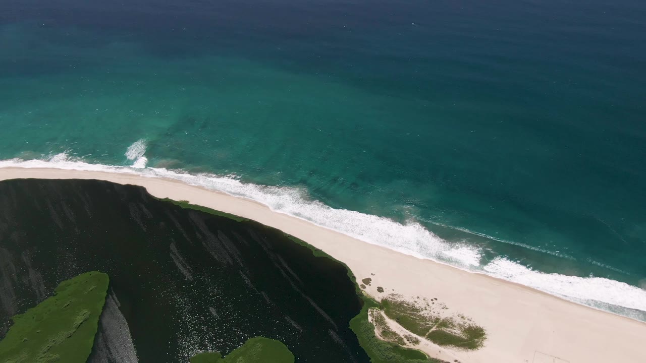 A second aerial moment over San Jose del Cabo shows the surf line spreading farther along the sand, with the grassy cliff occupying the lower left and no visible people.