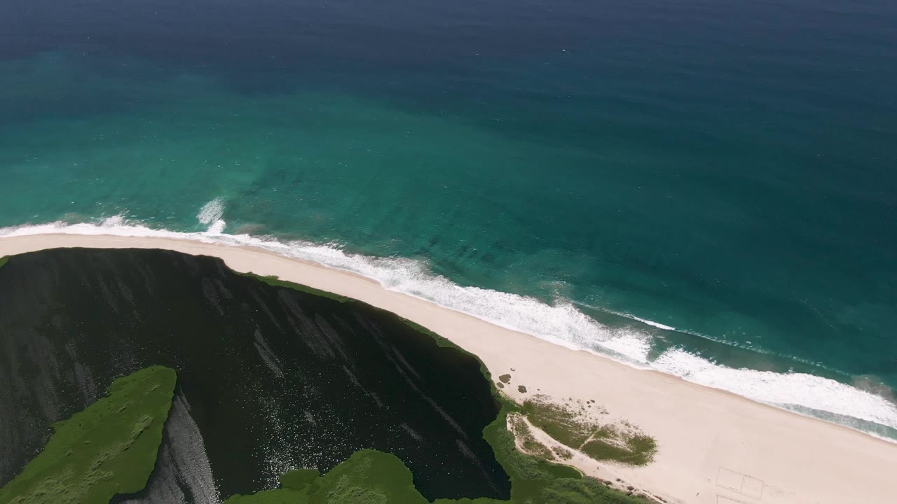 Aerial San Jose del Cabo coastline frame with turquoise water near shore, deeper blue ocean above, a pale beach, and the cliff edge lower in frame with no visible people.