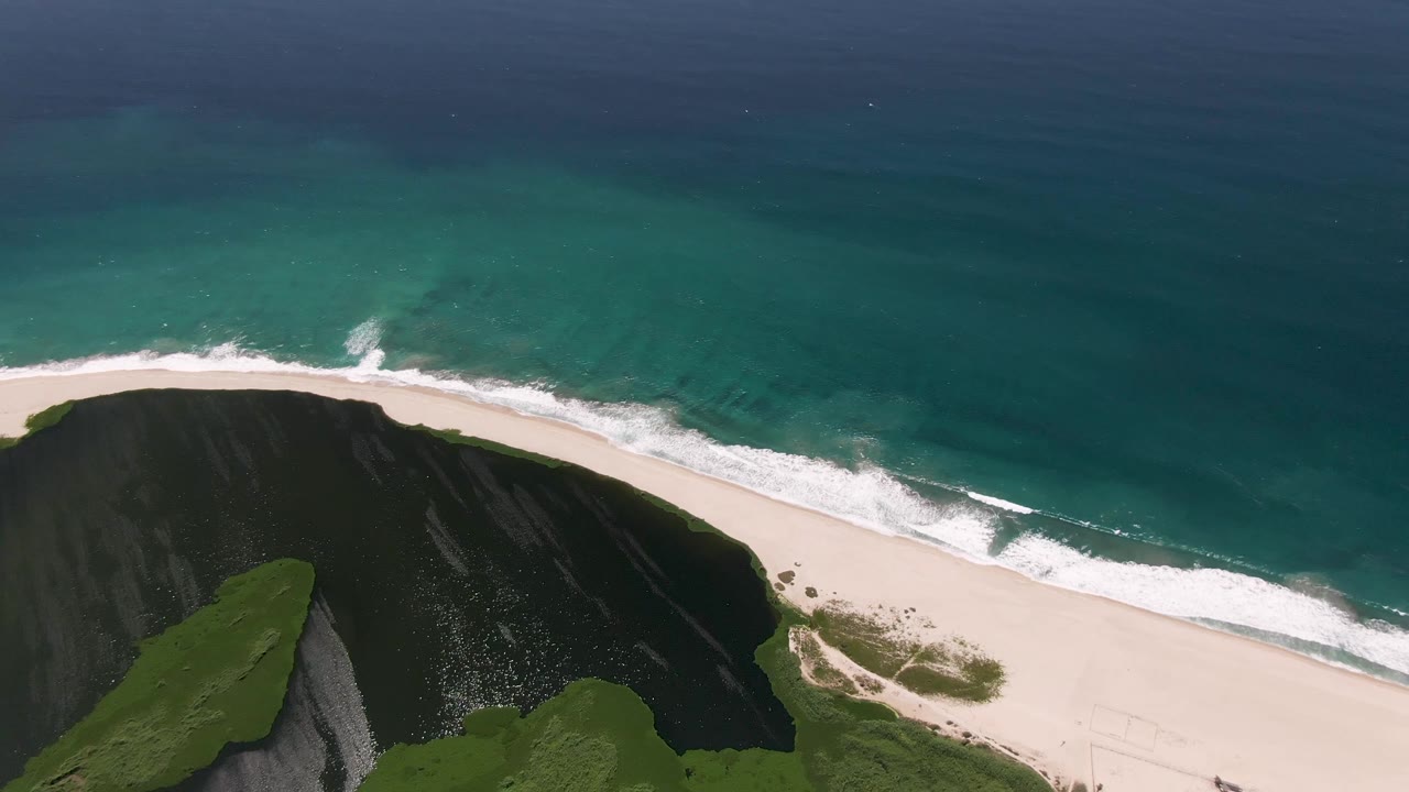 Aerial coastline with surf rolling onto sand beside a dark cliff.