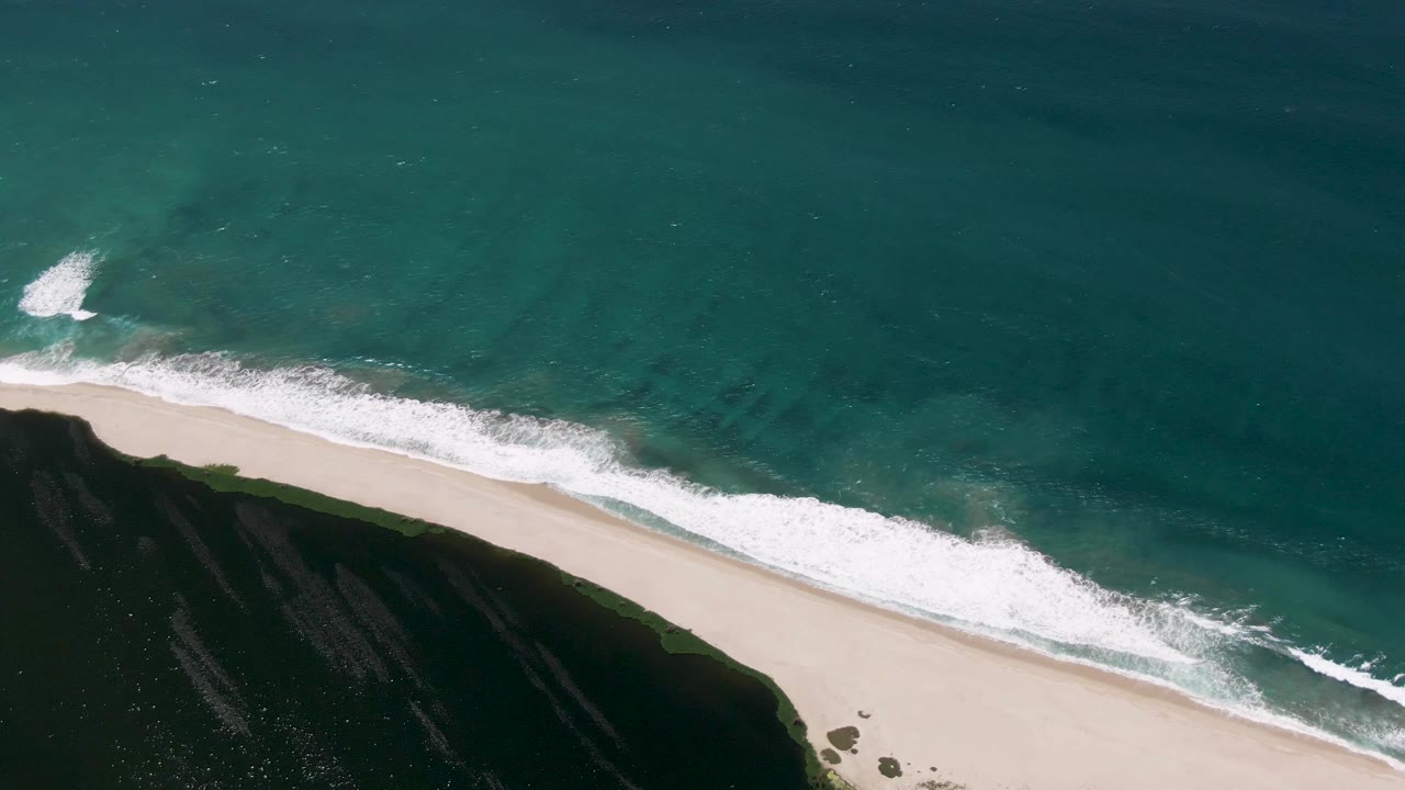 Aerial shoreline frame from San Jose Del Cabo, Mexico, where a wider band of white foam runs along the sand beside dark green coastal land, with no visible people.
