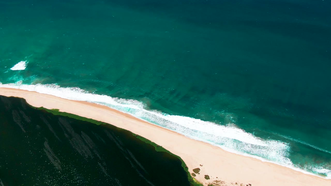 Overhead beach view in San Jose del Cabo, Mexico, with turquoise water, white surf, pale sand, a dark cliff edge, and no visible people at the start of the clip.