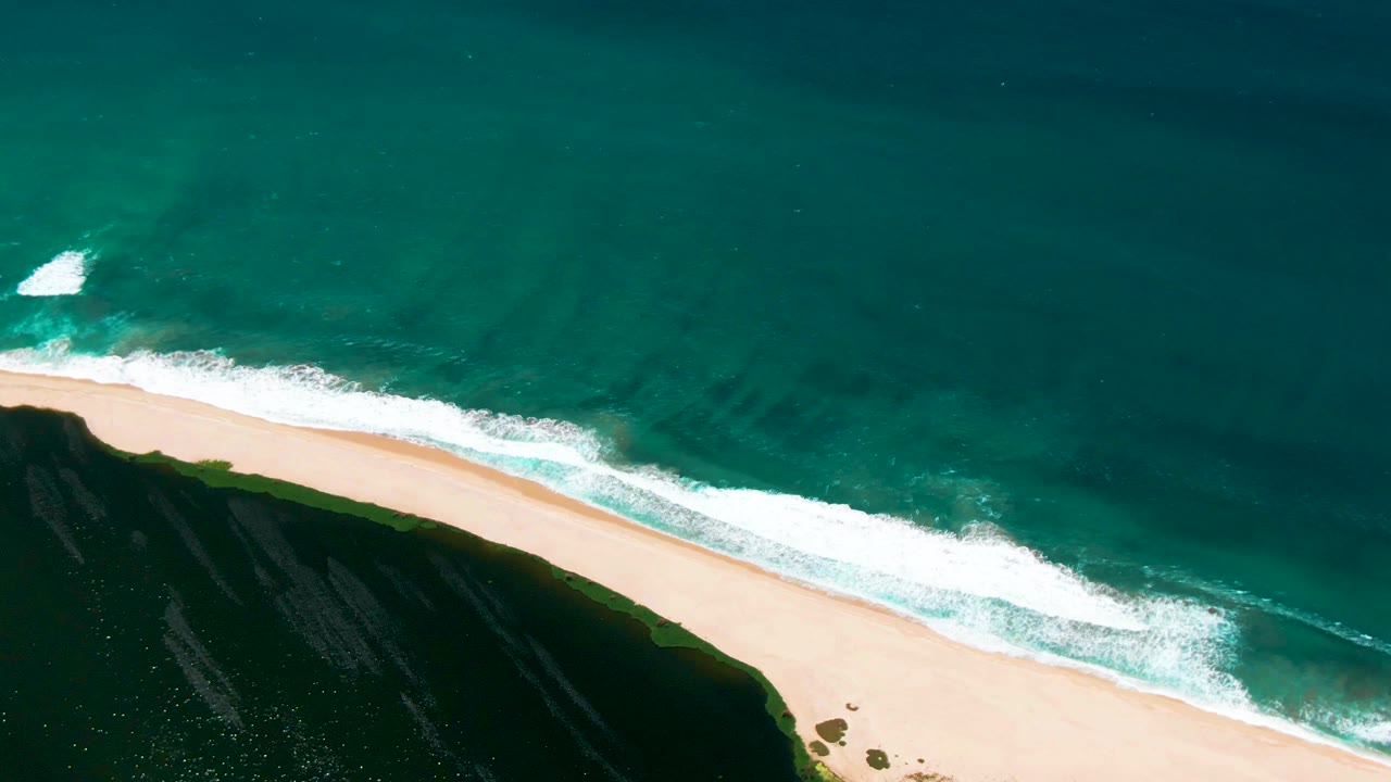 Aerial frame from San Jose del Cabo, Mexico, showing the white foam band slightly farther along the pale sand beside dark coastal rock, with no visible people.