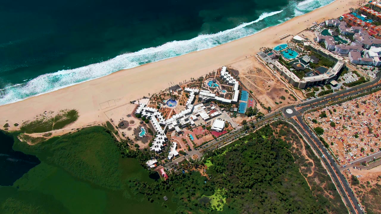 Aerial view of beach, resort buildings, road, greenery, and surf.