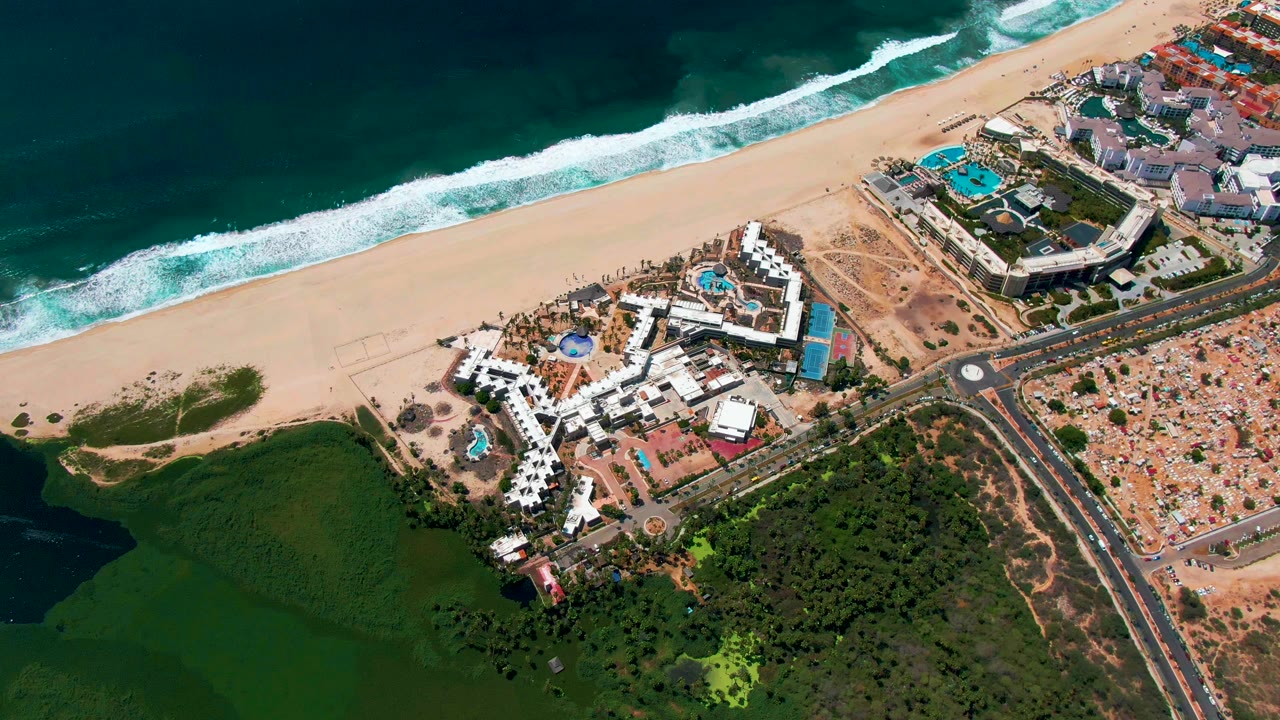 Aerial frame over San Jose Del Cabo showing the sandy beach and surf above resort buildings, a blue pool complex, road intersections, construction areas, and no visible people.