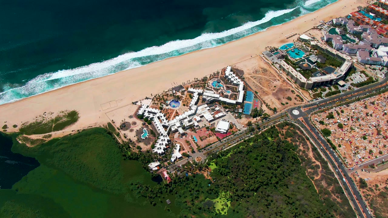 A high aerial frame in San Jose Del Cabo shows the beach and white surf across the top, clustered resort buildings and pools in the middle, dark green water and vegetation below, and no visible people.