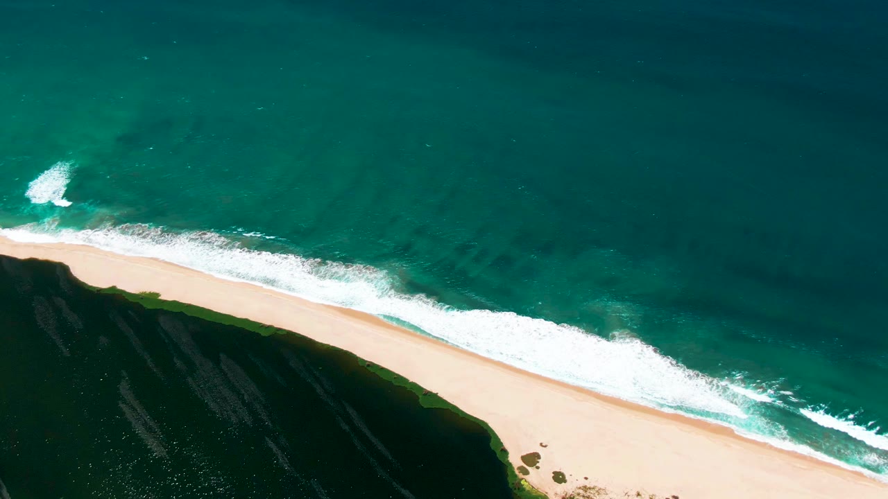 Aerial view of waves meeting a sandy beach beside a dark cliff.