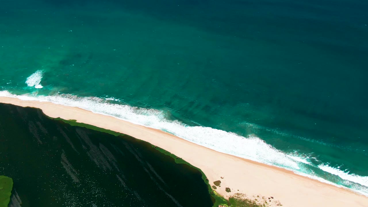 Aerial frame from San Jose del Cabo showing a broader foam band along the beach, deep green-blue water above it, dark cliff shadow at left, and no visible people.