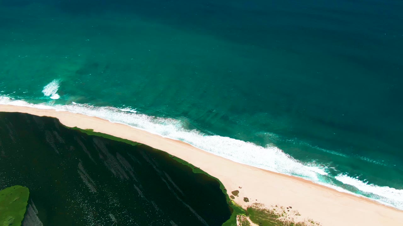 San Jose del Cabo aerial frame with the surf line shifted along the beach, darker ocean water toward the top, a pale sand strip through the middle, and no visible people.