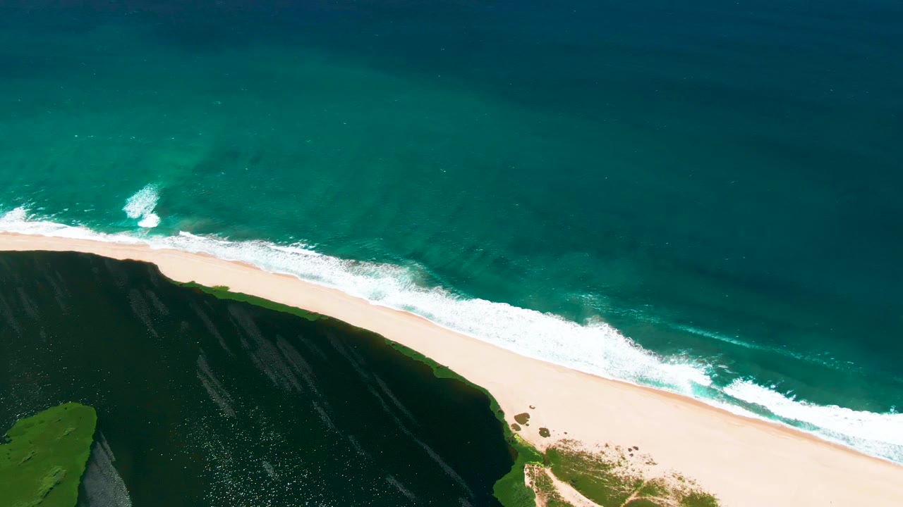 Mid-clip aerial view over San Jose del Cabo, where white foam runs nearly parallel to the sand, turquoise water fills most of the frame, and no visible people appear on the beach.