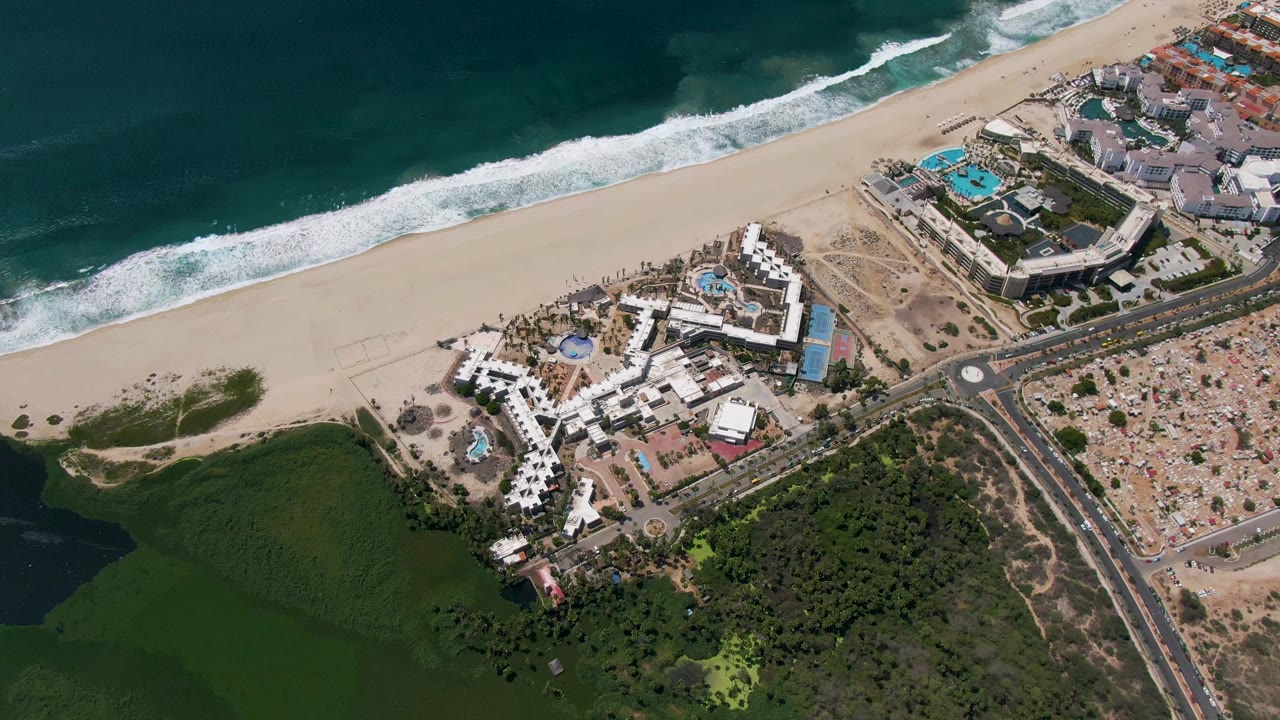 Aerial frame over San Jose del Cabo showing blue ocean, a wide sandy beach, resort buildings with pools, a coastal road, green vegetation, and no visible people.
