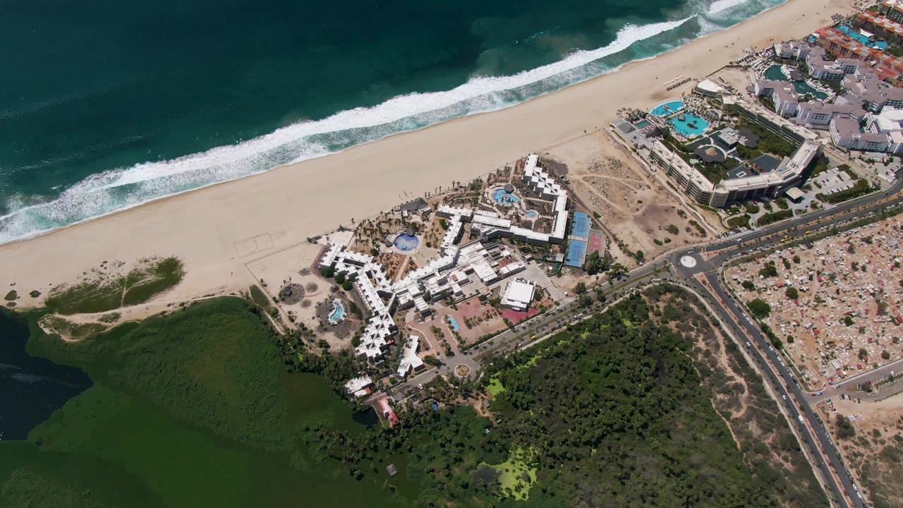 Aerial coastline with ocean waves, beach resorts, roads, and green terrain.