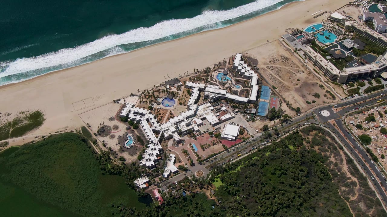 High aerial frame over a beachfront resort in San Jose del Cabo, Mexico, with white buildings, blue pools, a wide sandy beach, dark ocean water, roadways, and no visible people.