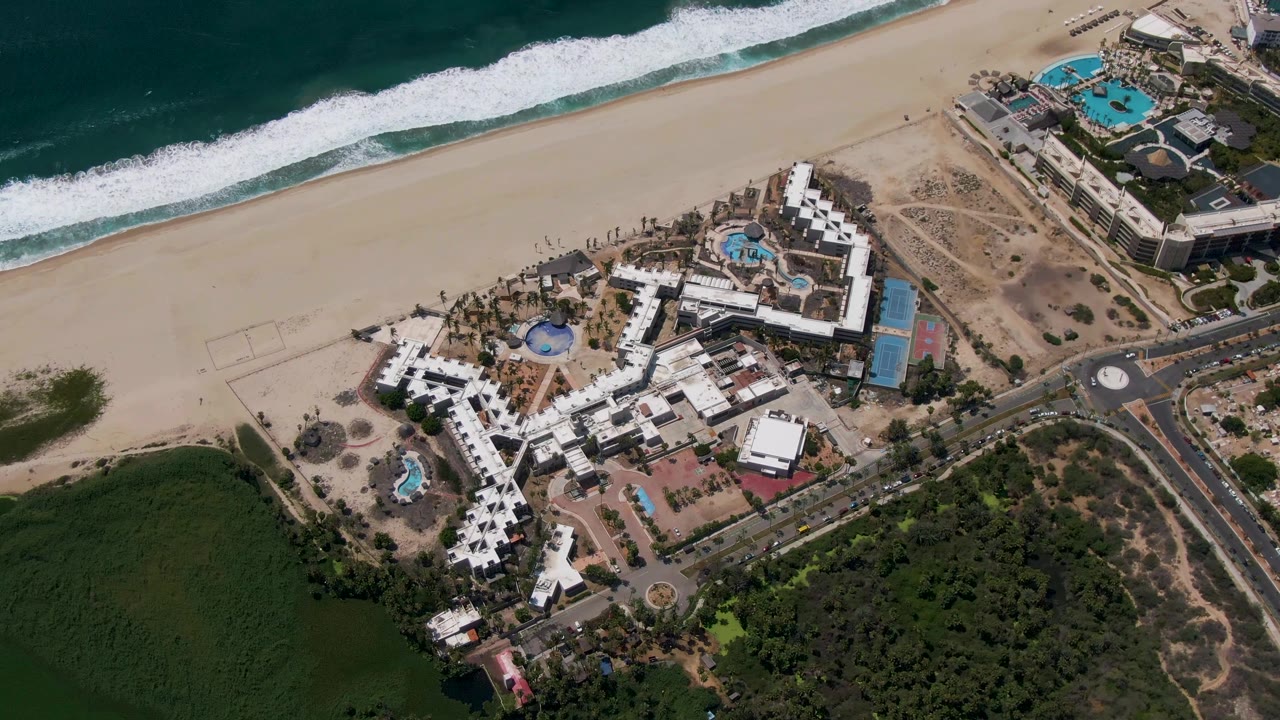 Aerial view in San Jose del Cabo, Mexico, showing the resort centered below the drone, with a diagonal shoreline, foamy waves, adjacent road, green vegetation, and no visible people.