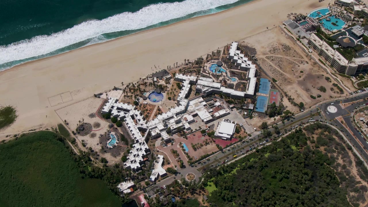 Overhead San Jose del Cabo resort frame with white buildings and blue pools below, surf at the upper edge, road and roundabout at the lower right, dry ground, and no visible people.