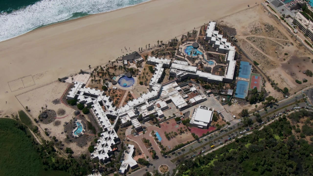 Second overhead frame of the San Jose Del Cabo resort shows the surf line shifted slightly along the beach, with pools and white roofs centered and no visible people.